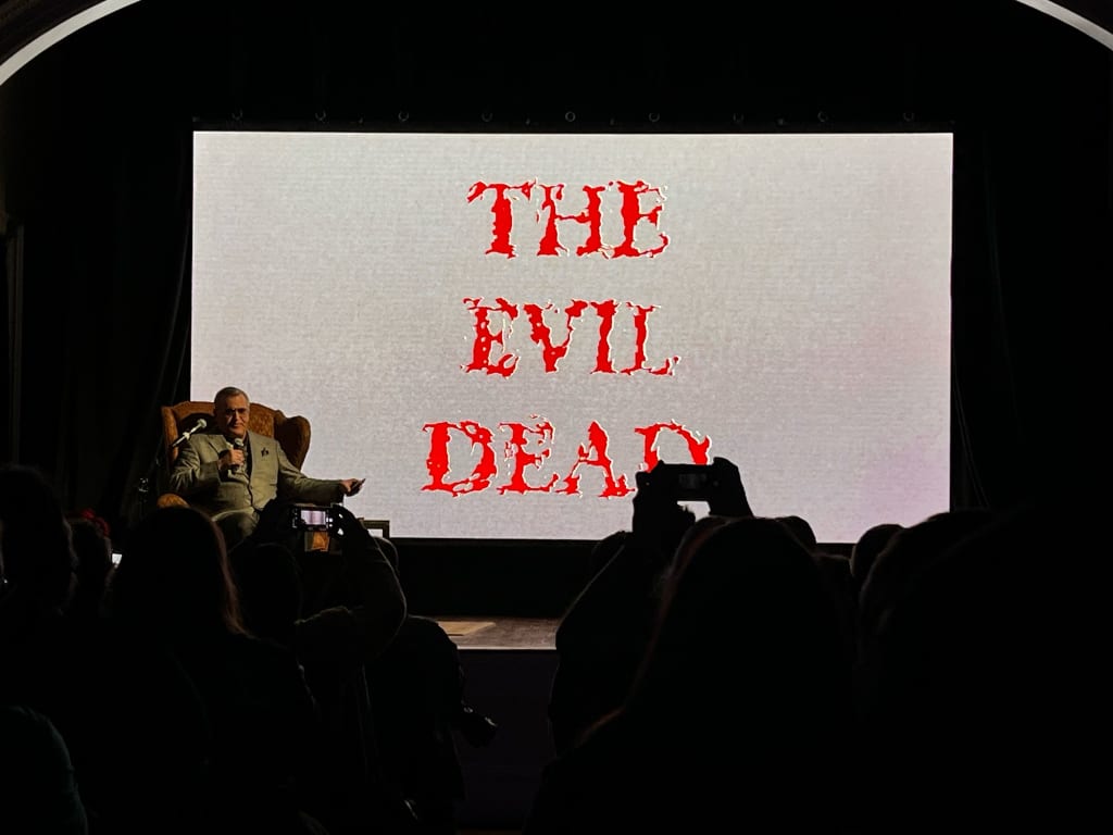 Bruce Campbell sits on a stage in a dark room with a large screen behind him that reads "The Evil Dead"